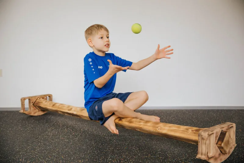 Junge sitzt im Spagat auf dem Balancierbalken und wirft einen Ball hoch
